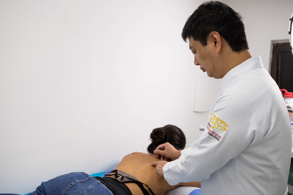 Dr. Marcus Yu Bin Pai, de jaleco, aplica agulhas na parte alta das costas de uma pessoa deitada de bruços, em sala de procedimento