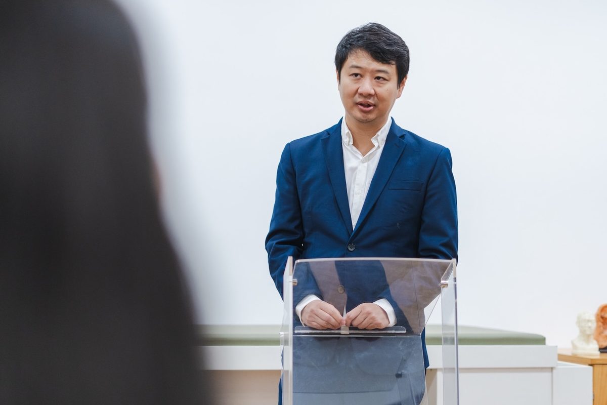 Dr. Marcus Yu Bin Pai fala atrás de um púlpito de acrílico, com uma ouvinte de costas em primeiro plano