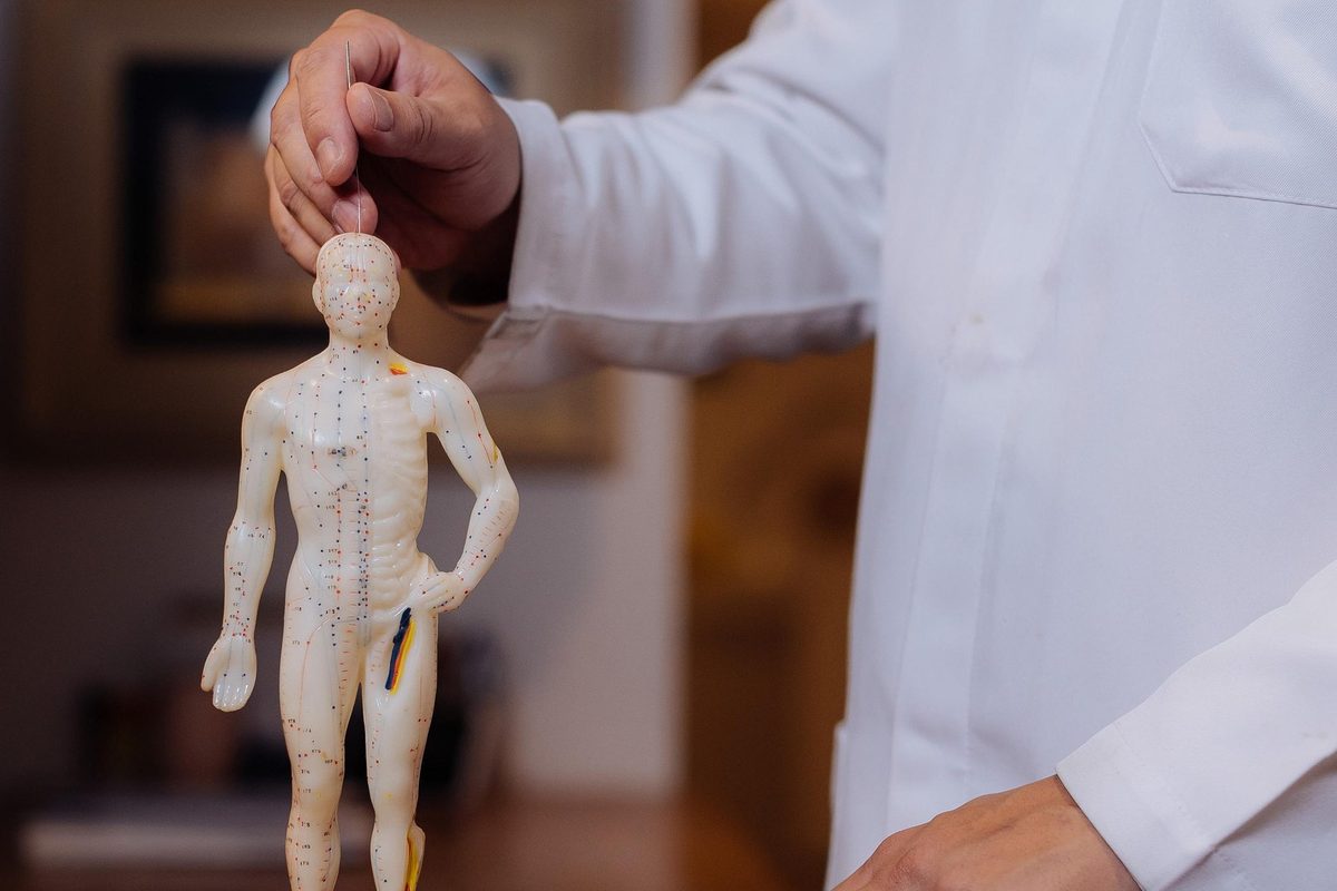 Mão do médico, de jaleco, segurando uma agulha sobre a cabeça de um boneco de acupuntura apoiado em um móvel de madeira