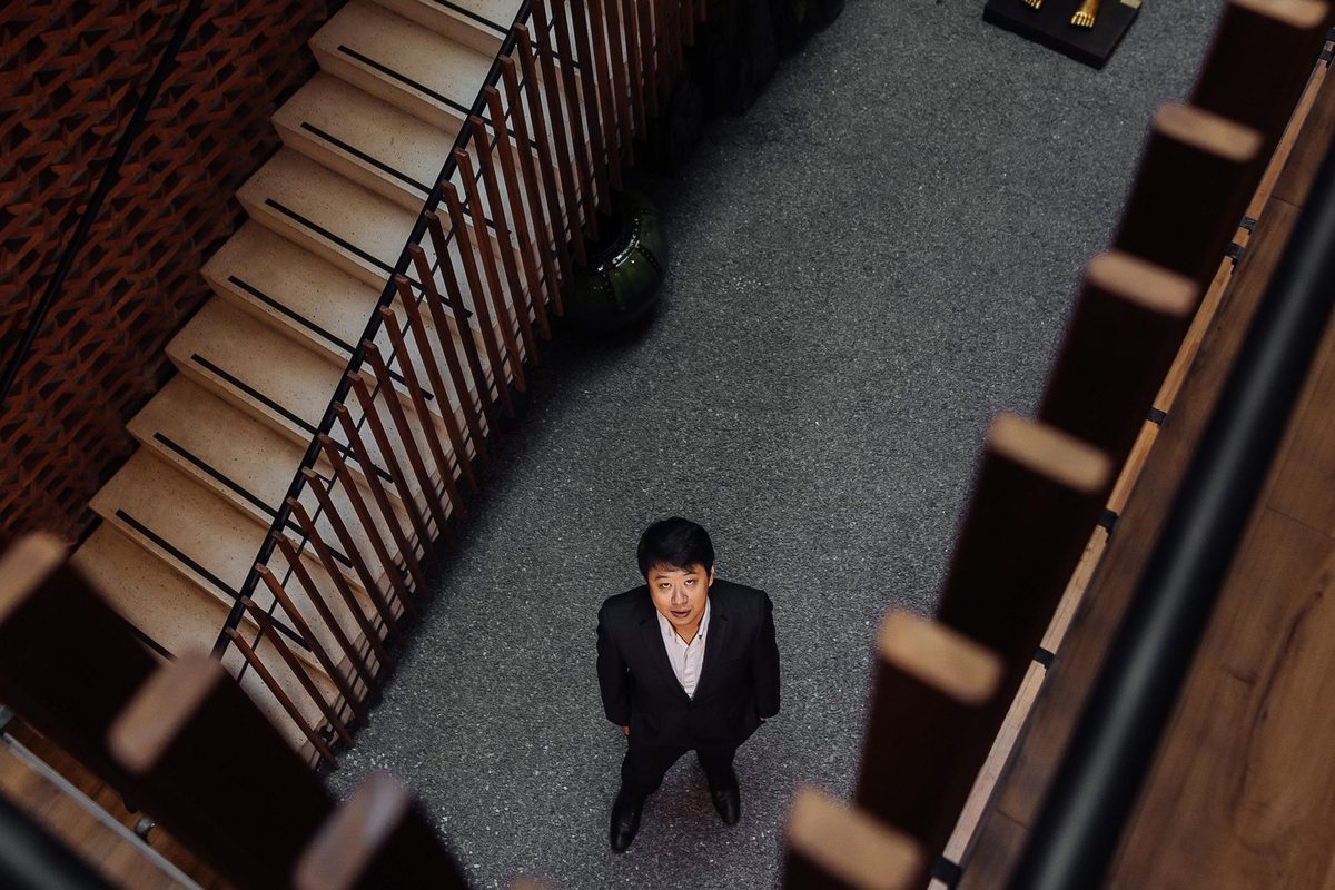 Átrio da clínica visto de cima, com a escada, os tijolos vazados e o ripado de madeira; Dr. Marcus Yu Bin Pai aparece pequeno, de pé no piso de pedrisco