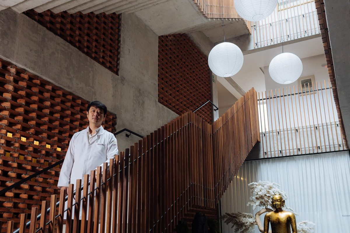 Dr. Marcus Yu Bin Pai de jaleco na escada do átrio da clínica, visto de longe sob a claraboia, com ripado de madeira, tijolos vazados e luminárias de papel