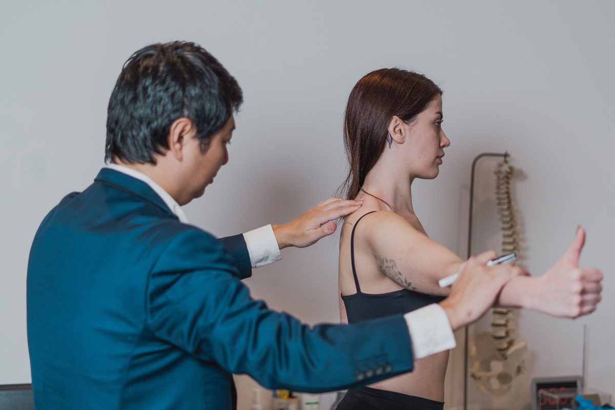 Dr. Marcus Yu Bin Pai palpa o ombro de uma pessoa que mantém o braço estendido à frente, em um teste do ombro