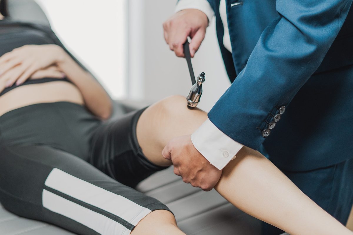 Mãos do médico aplicando o martelo de reflexos no joelho de uma pessoa deitada na maca