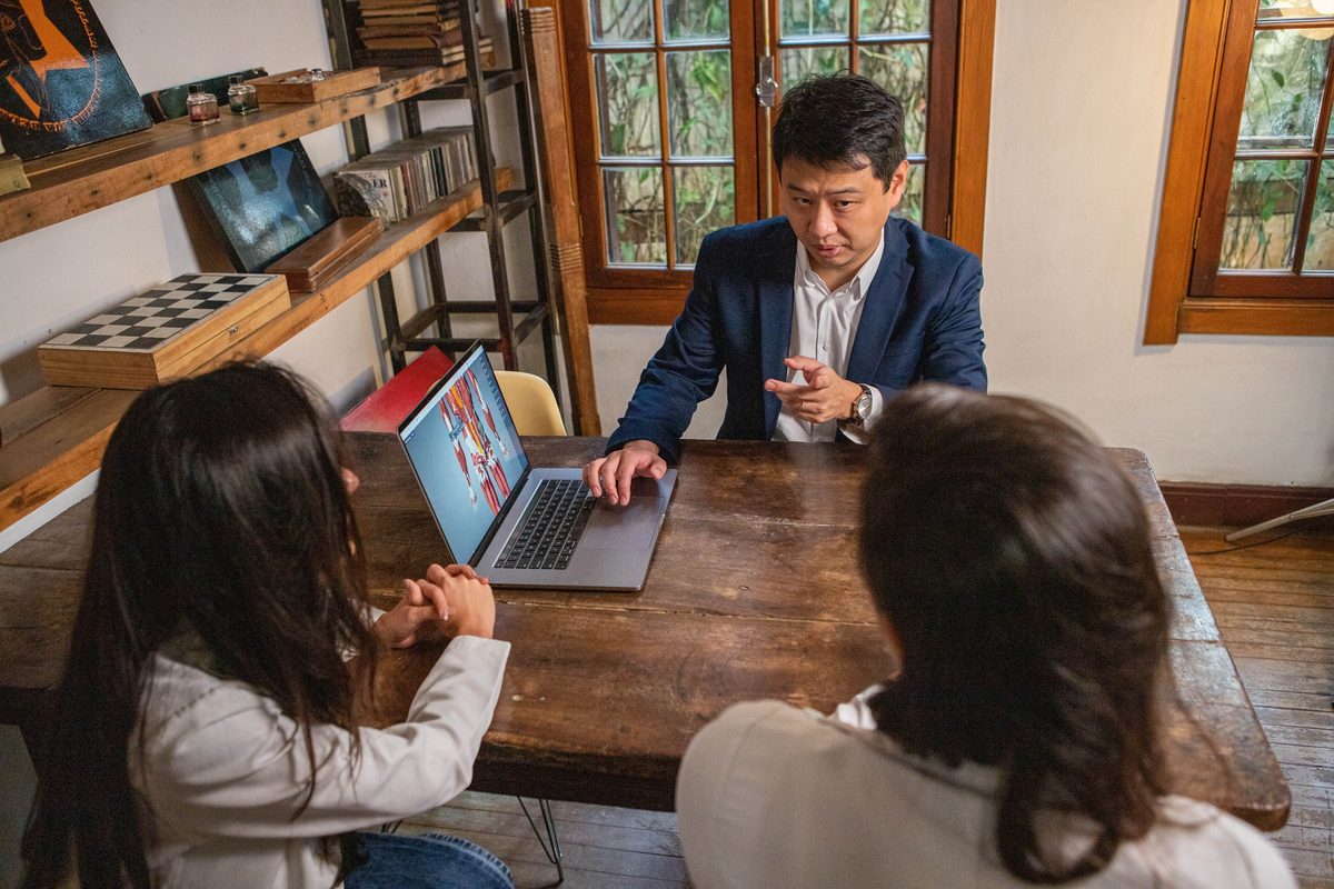 Dr. Marcus Yu Bin Pai, de blazer, explica um modelo anatômico na tela de um notebook a duas pessoas sentadas à mesa, vistas de costas