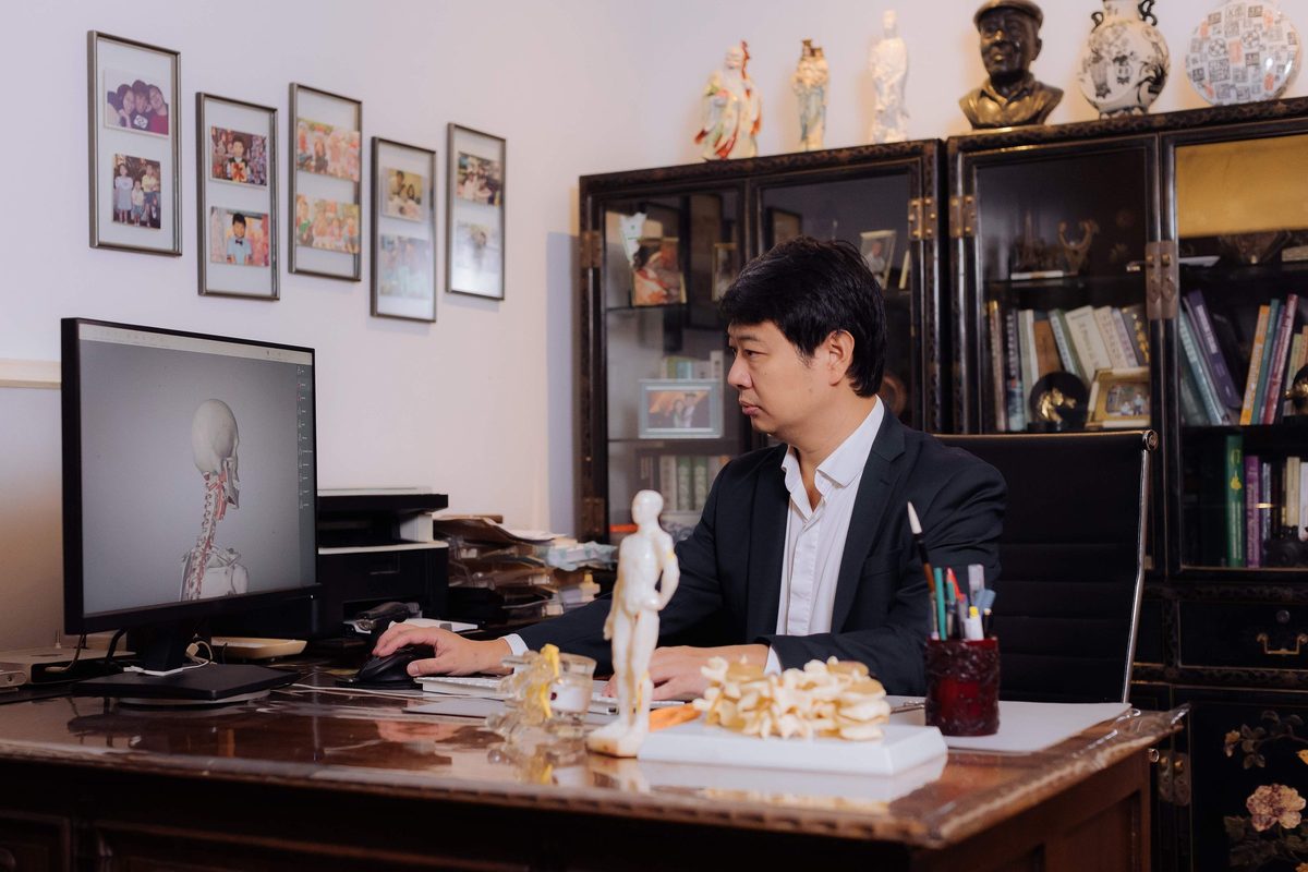Dr. Marcus Yu Bin Pai trabalha ao computador na mesa do consultório, com um modelo anatômico em 3D da cabeça e do pescoço na tela e modelos de coluna e de acupuntura sobre a mesa