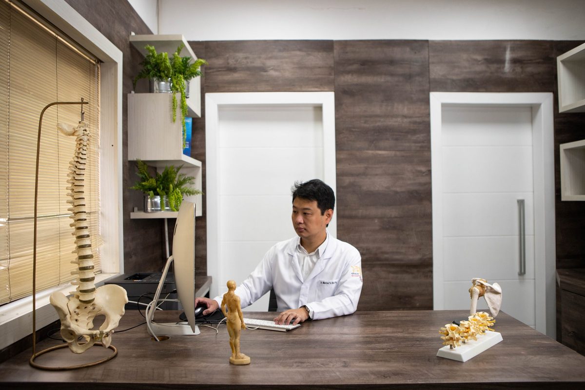 Dr. Marcus Yu Bin Pai de jaleco ao computador na mesa do consultório, com modelos de coluna e de ombro sobre a mesa