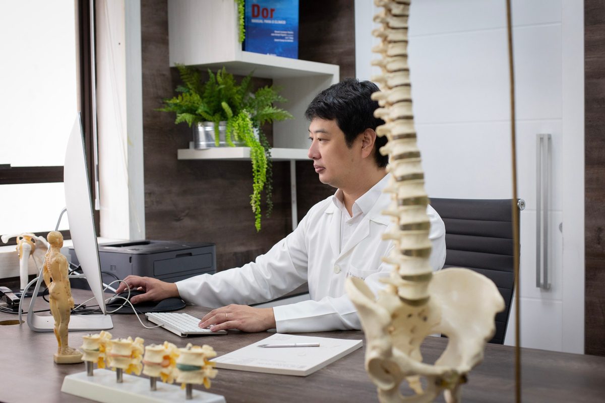Dr. Marcus Yu Bin Pai, de jaleco, trabalha ao computador na mesa do consultório, visto de perfil, com modelos de coluna vertebral em primeiro plano