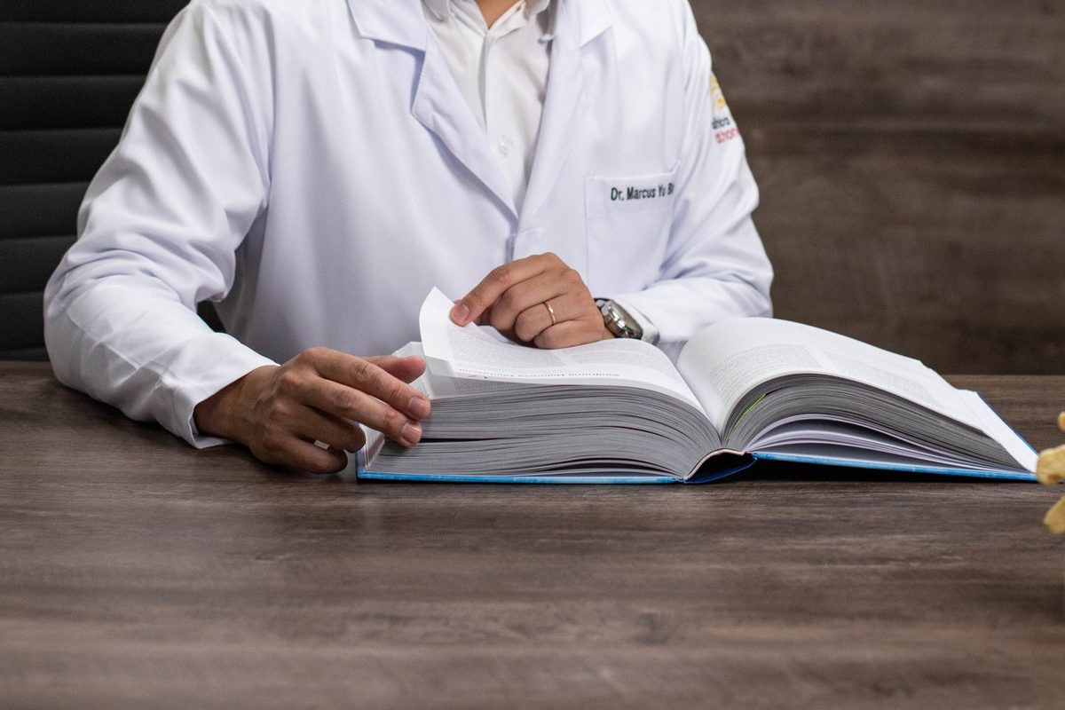 Mãos do médico, de jaleco, folheando um livro sobre a mesa do consultório