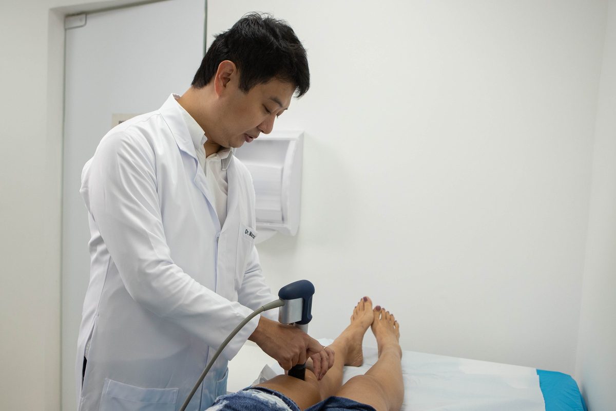 Dr. Marcus Yu Bin Pai, de jaleco, aplica terapia por ondas de choque no joelho de uma pessoa deitada na maca