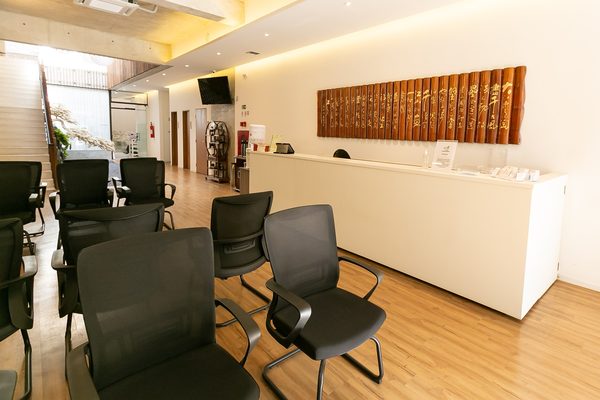 Balcão branco da recepção da clínica, com painel de bambu entalhado em caligrafia chinesa na parede e cadeiras da sala de espera em primeiro plano