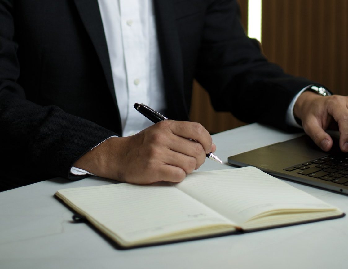 Mãos do Dr. Marcus Yu Bin Pai à mesa, com uma caneta sobre um caderno aberto, ao lado de um notebook