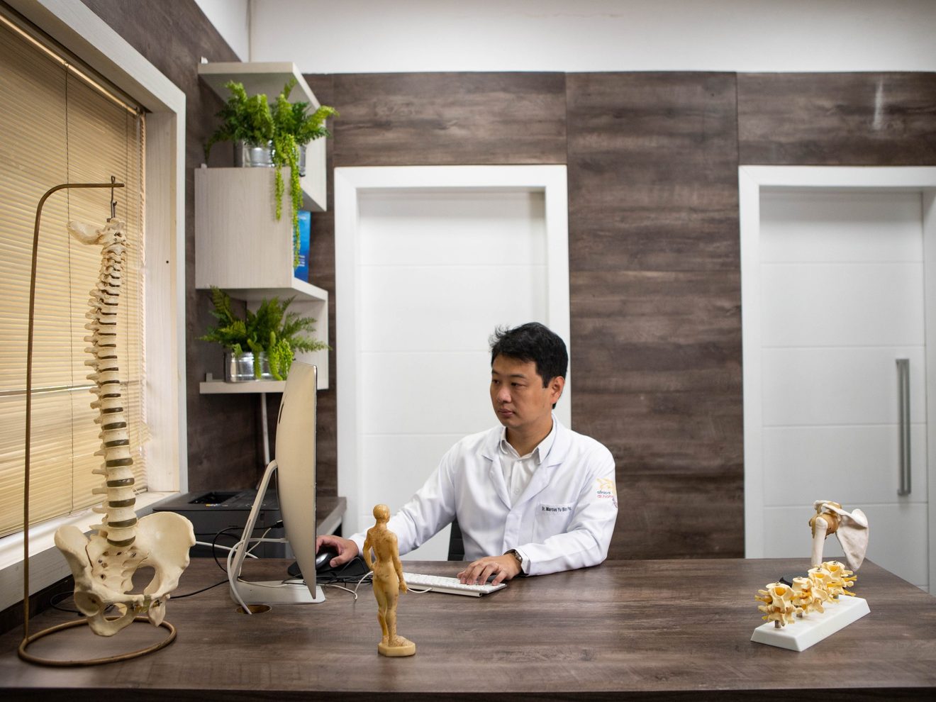 Dr. Marcus Yu Bin Pai de jaleco ao computador na mesa do consultório, com modelos de coluna e de ombro sobre a mesa