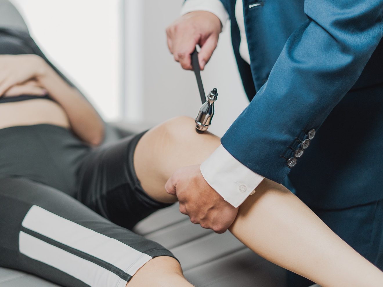 Mãos do médico aplicando o martelo de reflexos no joelho de uma pessoa deitada na maca