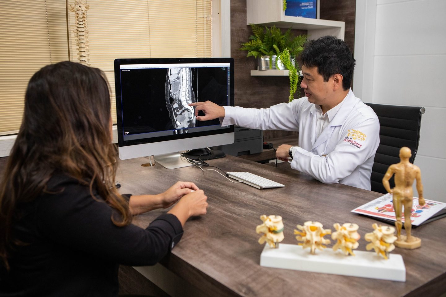 Dr. Marcus Yu Bin Pai aponta no monitor uma imagem de exame da coluna lombar para uma pessoa sentada à sua frente, vista de costas