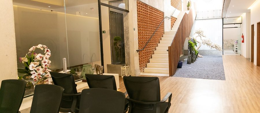 Sala de espera da clínica com vista para o átrio: escada, parede de tijolos vazados, ripado de madeira e jardim de pedras ao fundo