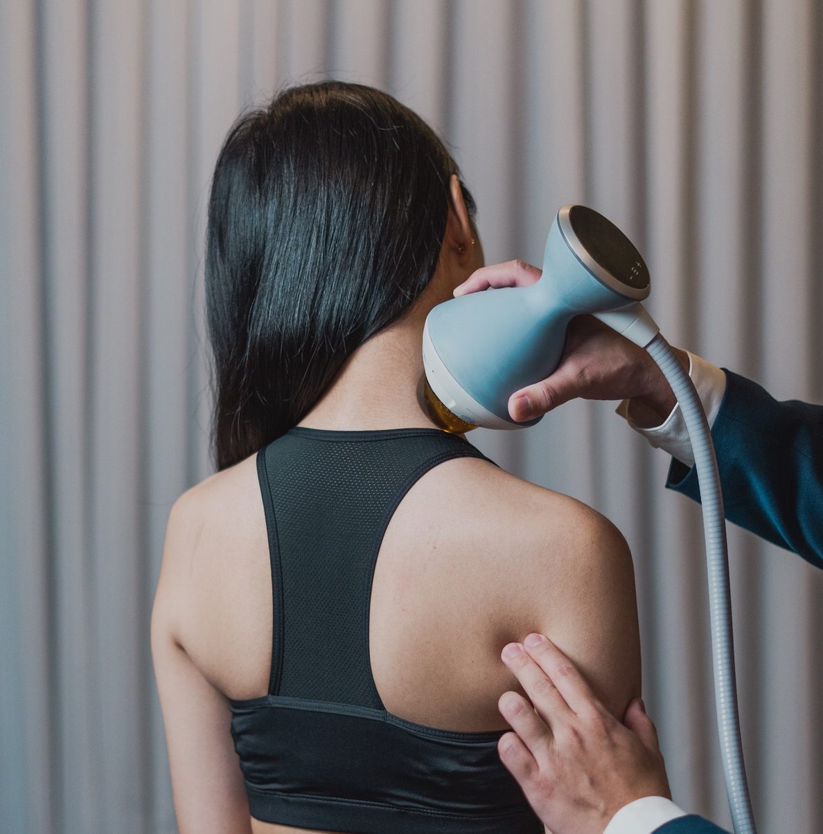 Aplicação de terapia por ondas de choque focal na região cervical de uma pessoa vista de costas; do médico aparecem apenas os braços e as mãos