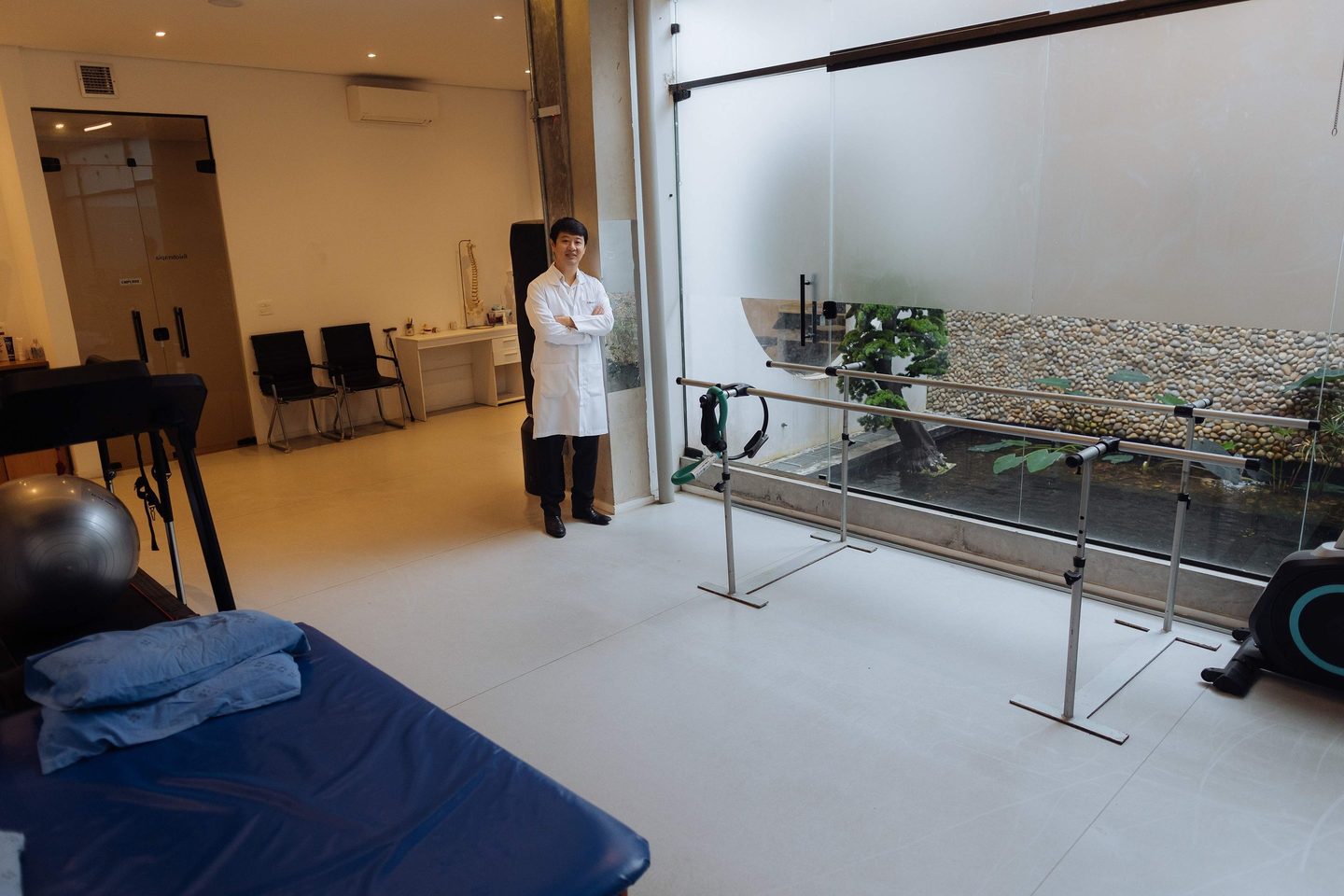 Sala de reabilitação da clínica com barras paralelas e vista para a parede de seixos atrás do vidro; Dr. Marcus Yu Bin Pai de jaleco aparece ao fundo