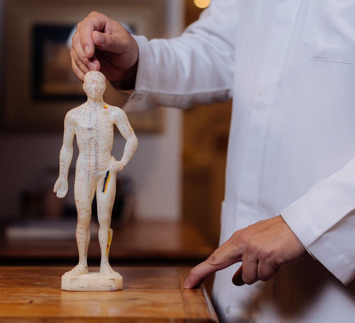 Mão do médico posiciona uma agulha na cabeça de um boneco de acupuntura sobre um móvel de madeira