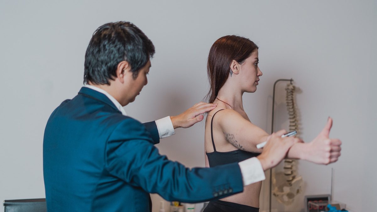 Dr. Marcus Yu Bin Pai palpa o ombro de uma pessoa que mantém o braço estendido à frente, durante o exame do ombro