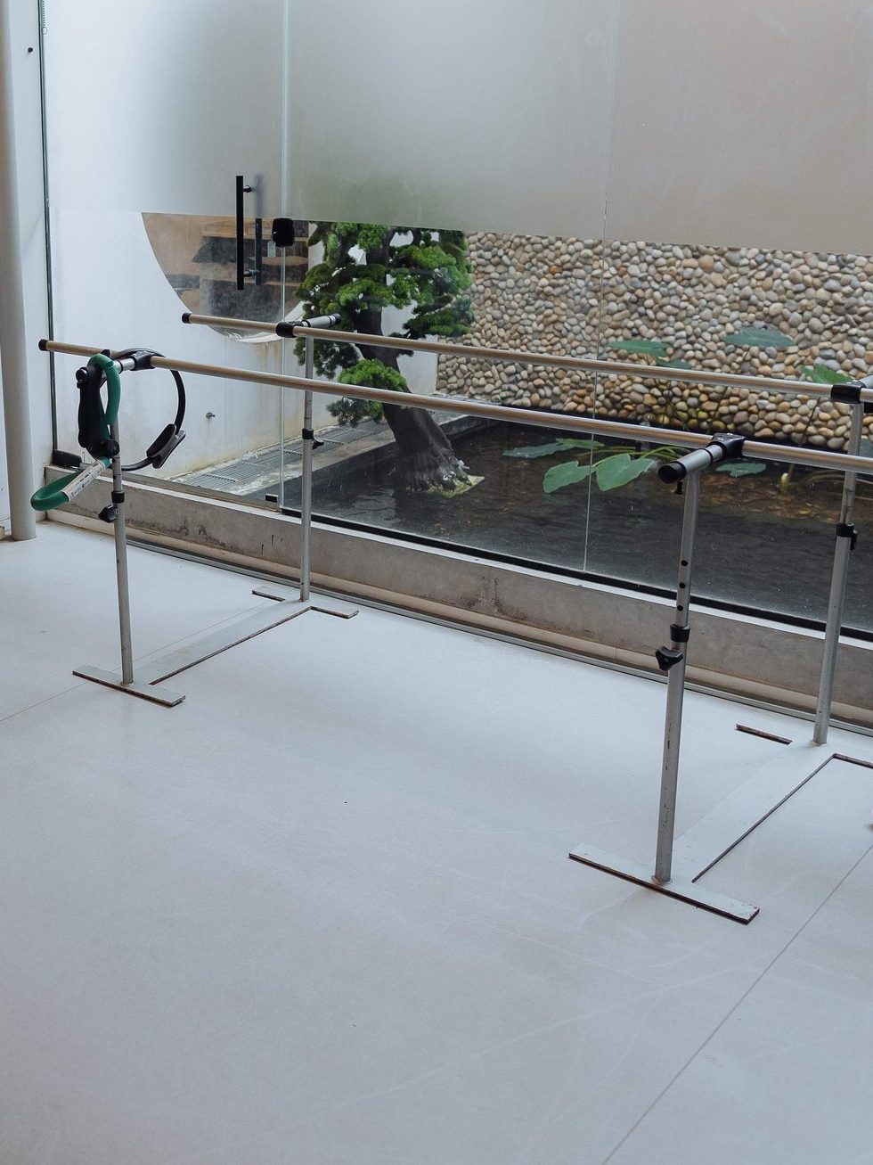 Barras paralelas da sala de reabilitação da clínica, diante do painel de vidro e do jardim interno com parede de seixos