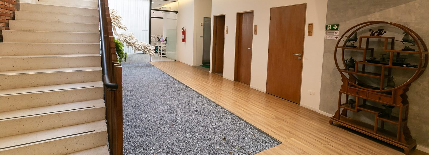 Térreo do átrio da clínica, com escada, piso de pedrisco, portas dos consultórios e estante chinesa de madeira