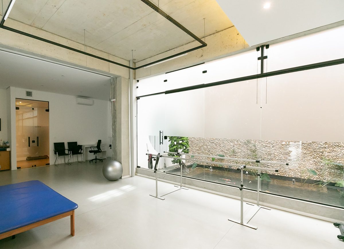 Sala de reabilitação da clínica com tablado azul, bola de exercício, barras paralelas e painel de vidro com vista para o jardim de seixos