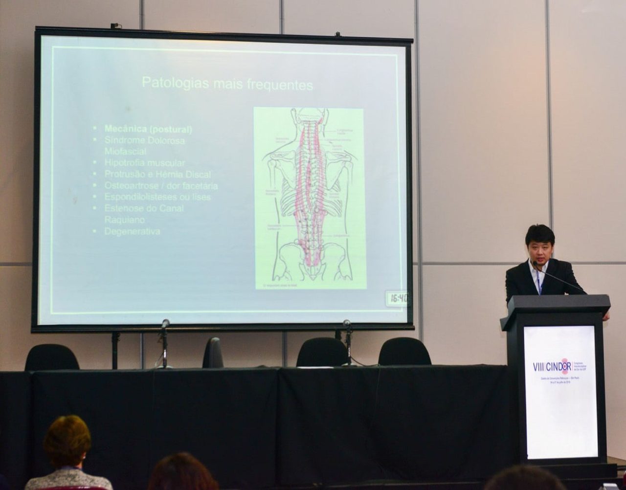 Dr. Marcus Yu Bin Pai fala no púlpito do VIII CINDOR, com slide sobre as patologias mais frequentes da coluna projetado no telão