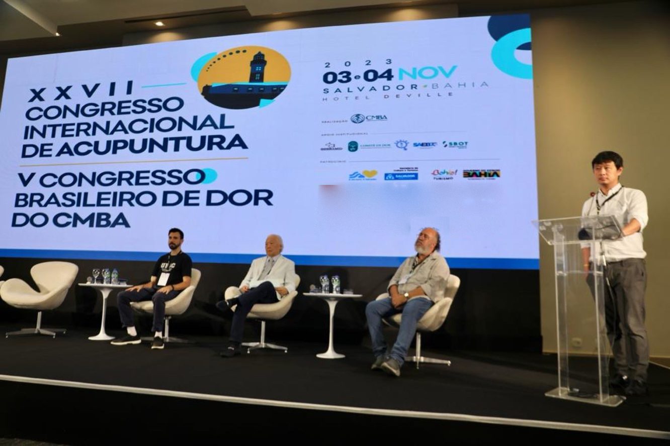 Palco do XXVII Congresso Internacional de Acupuntura e V Congresso Brasileiro de Dor do CMBA, em Salvador: uma pessoa ao púlpito de acrílico e três pessoas sentadas; o Dr. Marcus Yu Bin Pai está entre as quatro