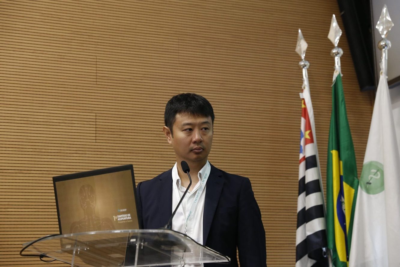 Dr. Marcus Yu Bin Pai ao púlpito de acrílico, como moderador do 1º Simpósio de Acupuntura do Cremesp, com bandeiras ao lado