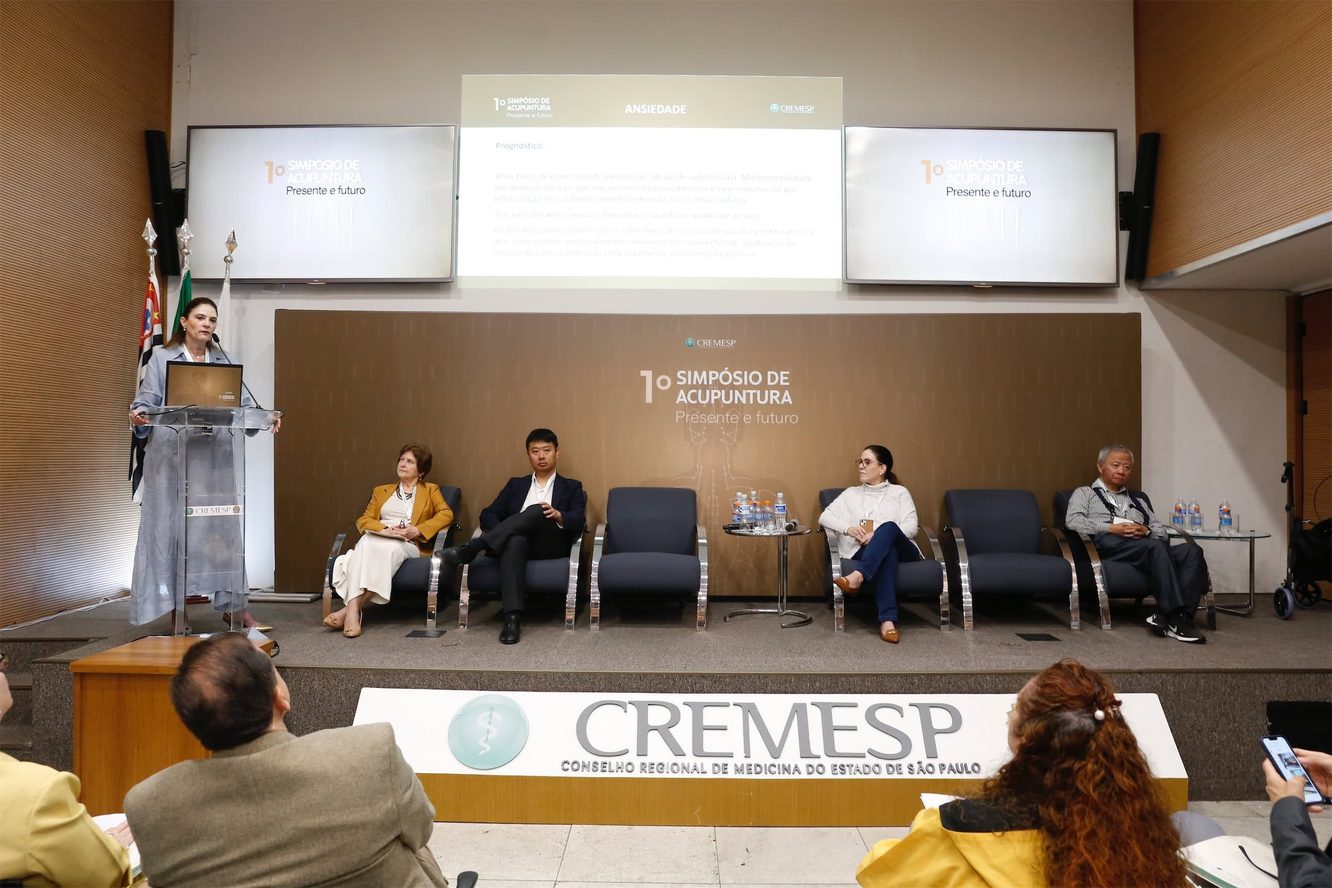 Mesa do 1º Simpósio de Acupuntura do Cremesp: uma pessoa fala ao púlpito e quatro pessoas estão sentadas no palco, entre elas o Dr. Marcus Yu Bin Pai, com a plateia de costas em primeiro plano