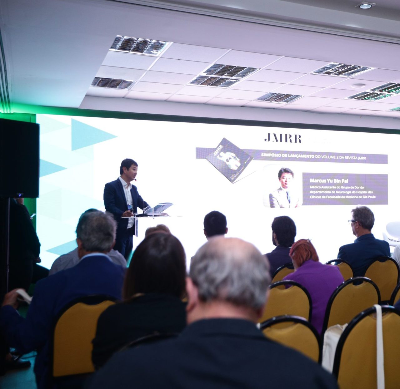 Dr. Marcus Yu Bin Pai fala no simpósio de lançamento da revista JMRR, com a plateia de costas e o slide com seu nome ao fundo
