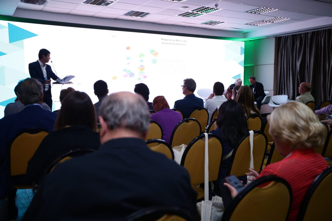 Dr. Marcus Yu Bin Pai apresenta o mapa de evidência da acupuntura para dor em um painel de LED, visto do fundo da sala, com a plateia de costas
