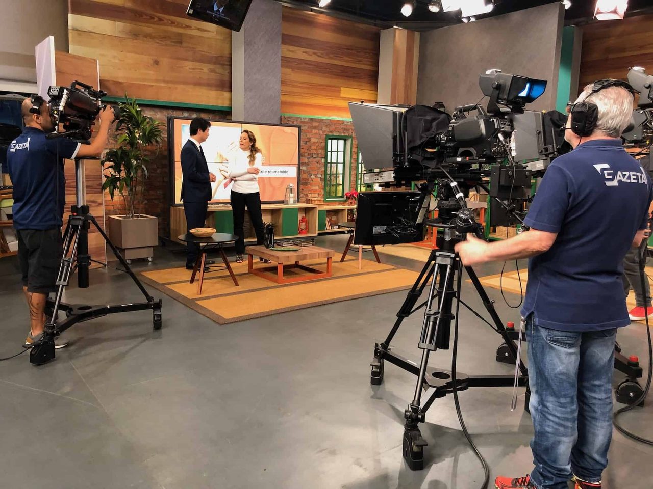 Bastidores de gravação do programa Mulheres em estúdio de televisão: duas pessoas conversam diante de um telão, entre elas o Dr. Marcus Yu Bin Pai, com câmeras e as pessoas que as operam em primeiro plano