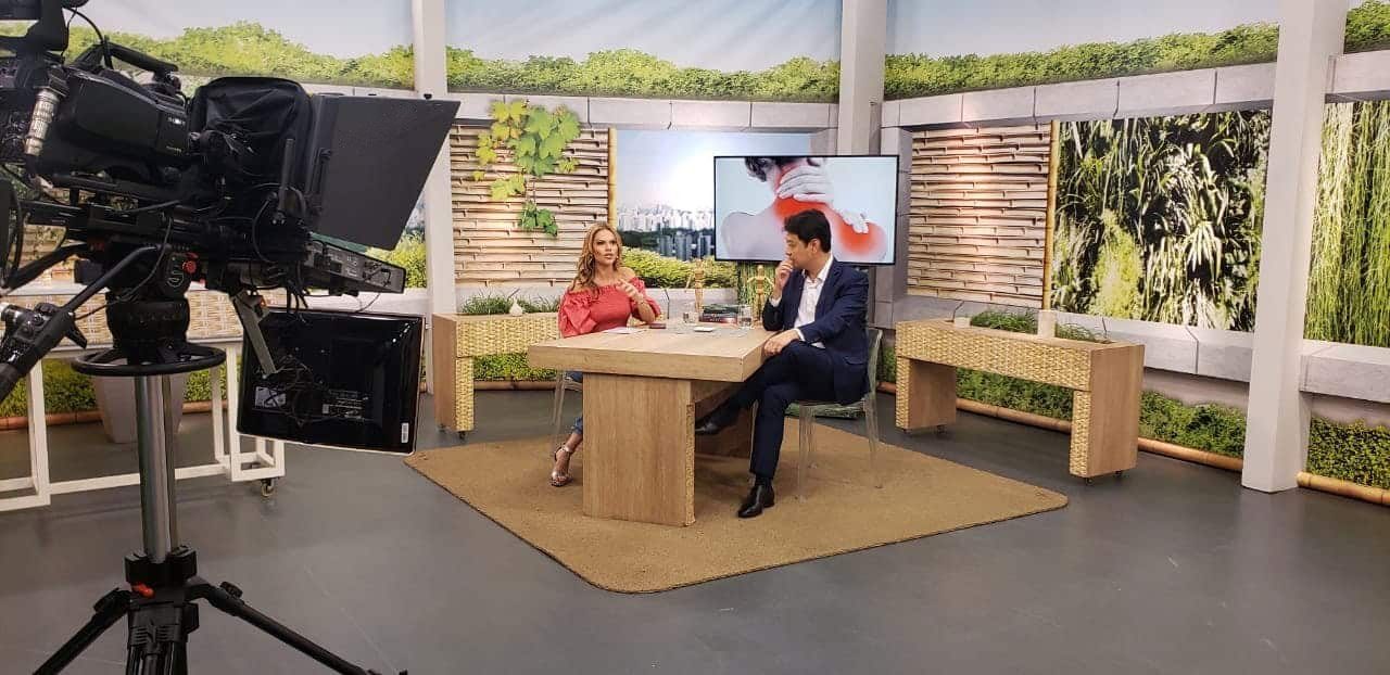 Entrevista no programa Você Bonita: o Dr. Marcus Yu Bin Pai e outra pessoa conversam sentados à mesa do cenário, vistos de longe por trás de uma câmera com teleprompter