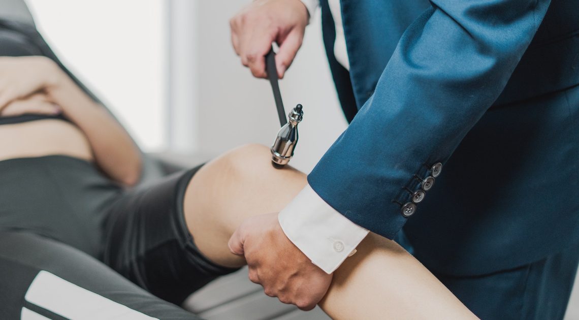 Mãos do médico aplicando o martelo de reflexos no joelho de uma pessoa deitada na maca