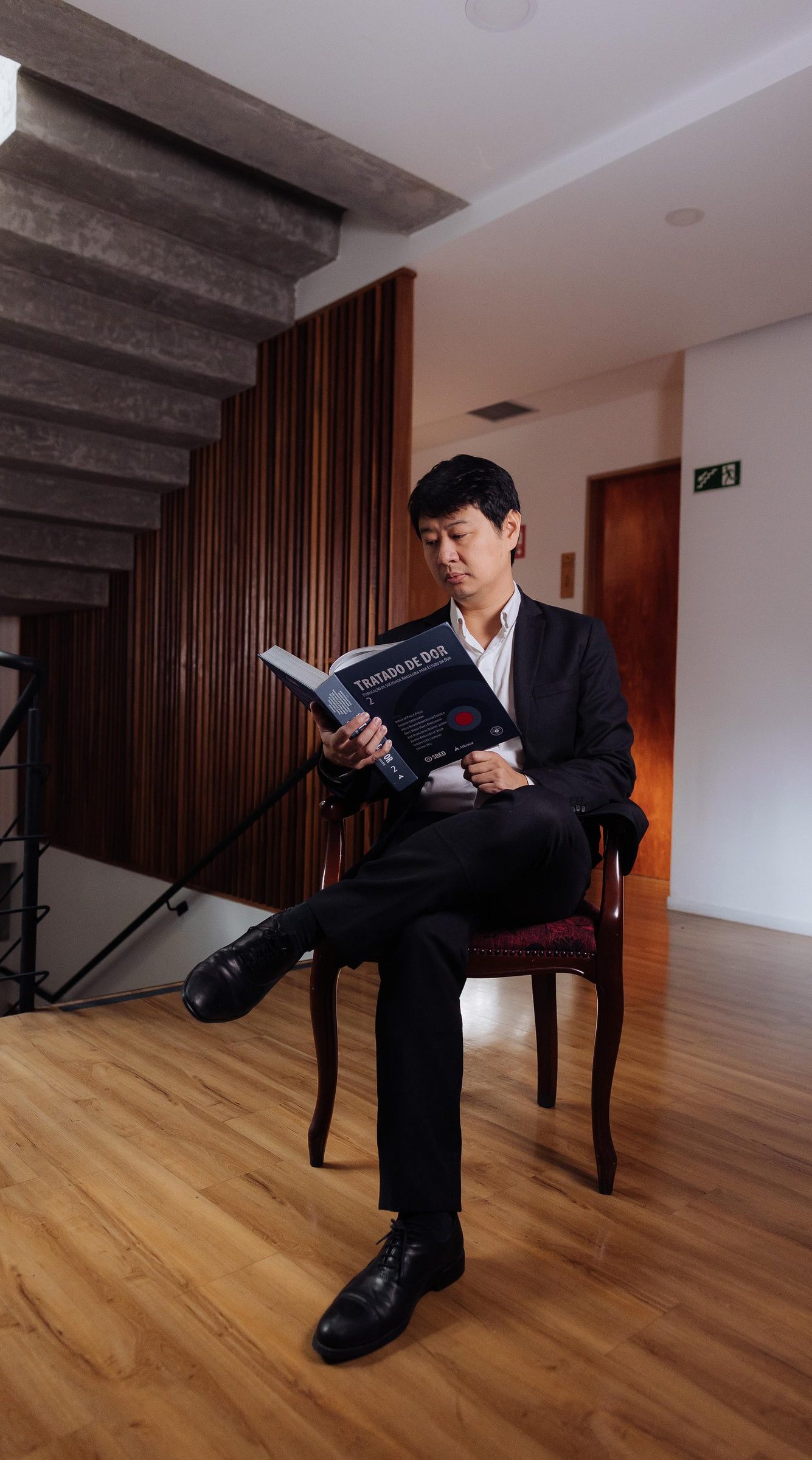 Dr. Marcus Yu Bin Pai sentado em uma cadeira, lendo o livro Tratado de Dor, ao lado da escada de concreto e do painel de ripas de madeira