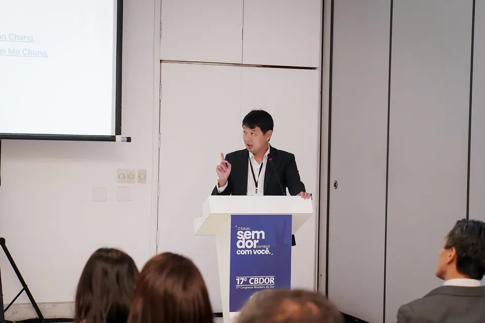 Dr. Marcus Yu Bin Pai gesticula ao púlpito do 17º Congresso Brasileiro de Dor, com a plateia de costas em primeiro plano