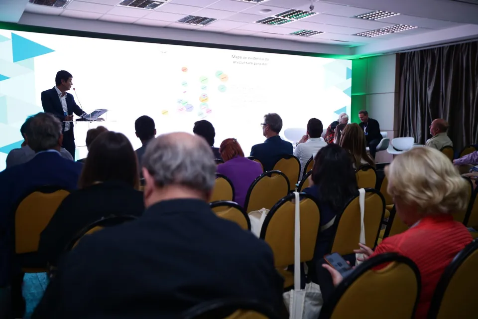 Dr. Marcus Yu Bin Pai apresenta o mapa de evidência da acupuntura para dor em um painel de LED, visto do fundo da sala, com a plateia de costas