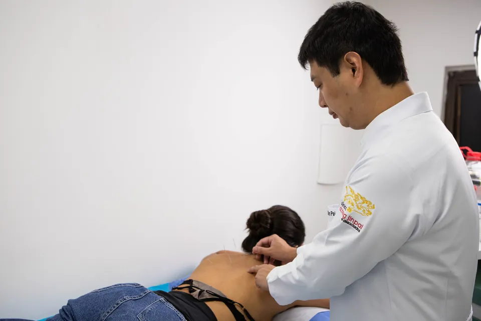 Dr. Marcus Yu Bin Pai de jaleco aplica agulhas na parte alta das costas de uma pessoa deitada de bruços