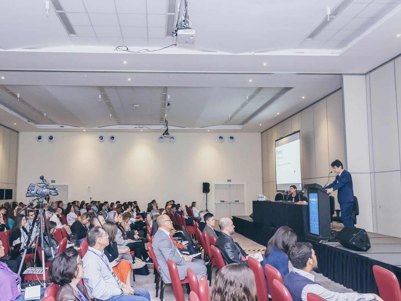 Dr. Marcus Yu Bin Pai fala ao púlpito de um auditório cheio durante congresso de dor, visto do fundo da sala