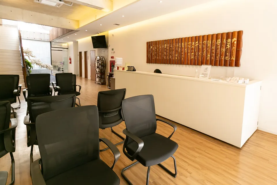 Balcão branco da recepção da clínica, com painel de bambu entalhado em caligrafia chinesa na parede e cadeiras da sala de espera em primeiro plano