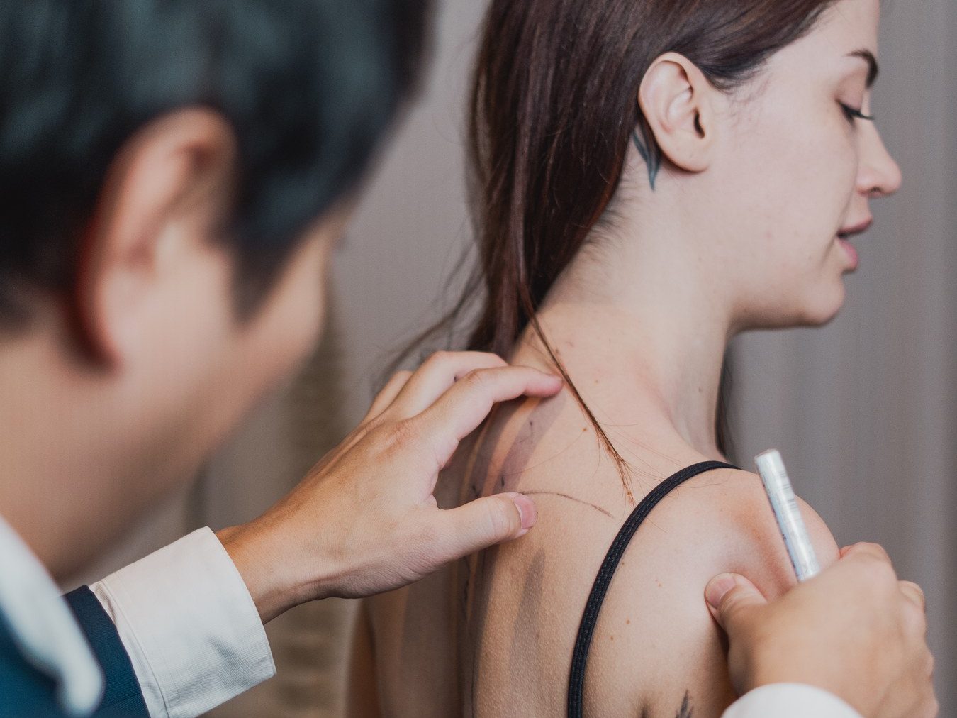 O médico, de costas e desfocado, palpa a região do pescoço e do ombro de uma pessoa e segura uma caneta de marcação