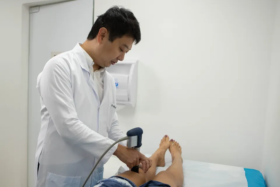 Dr. Marcus Yu Bin Pai de jaleco aplica terapia por ondas de choque no joelho de uma pessoa deitada na maca